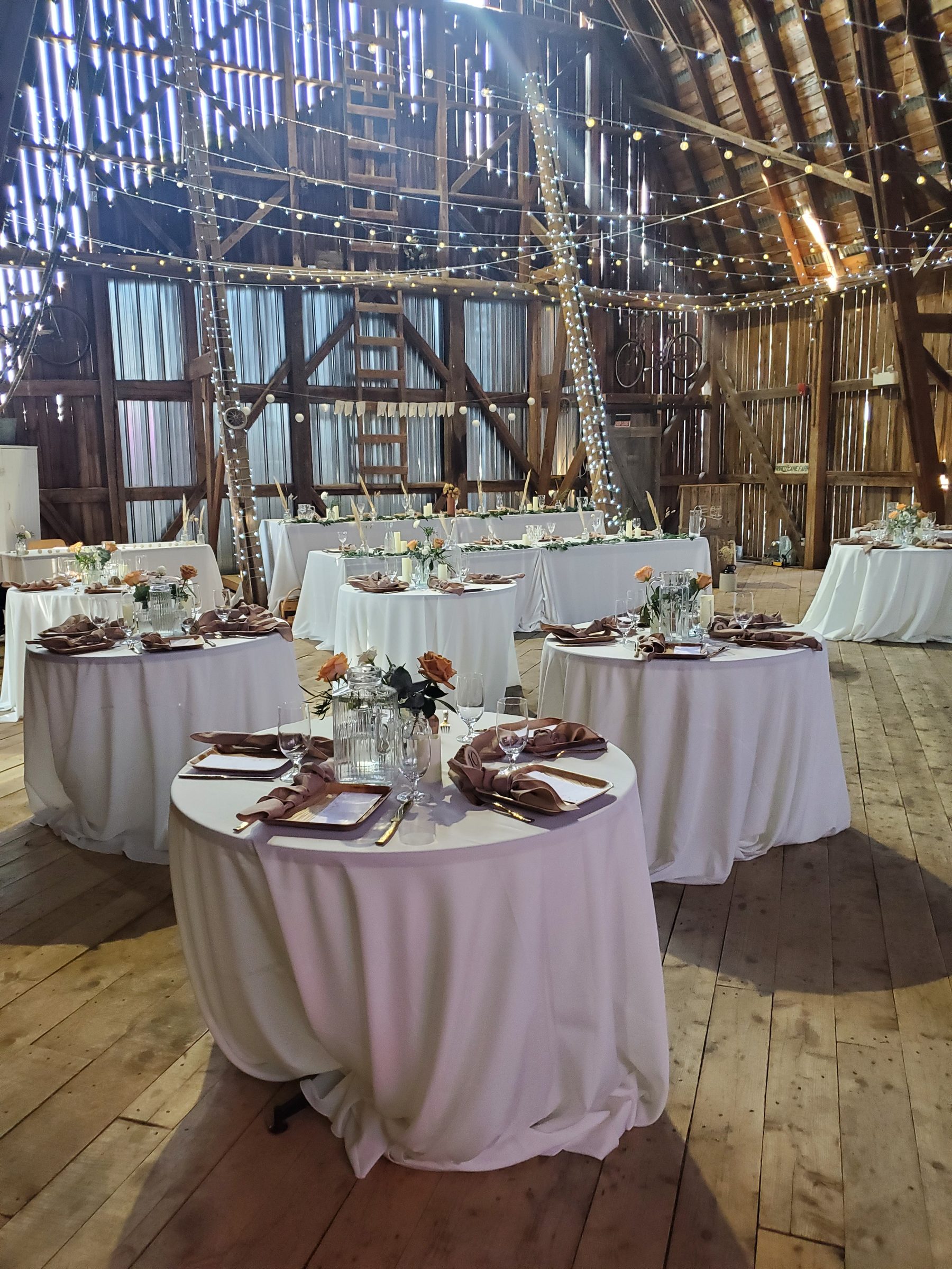Barn interior set for a wedding reception with round tables, dusty rose napkins, a long head table, and a canopy of string lights beneath the timber roof