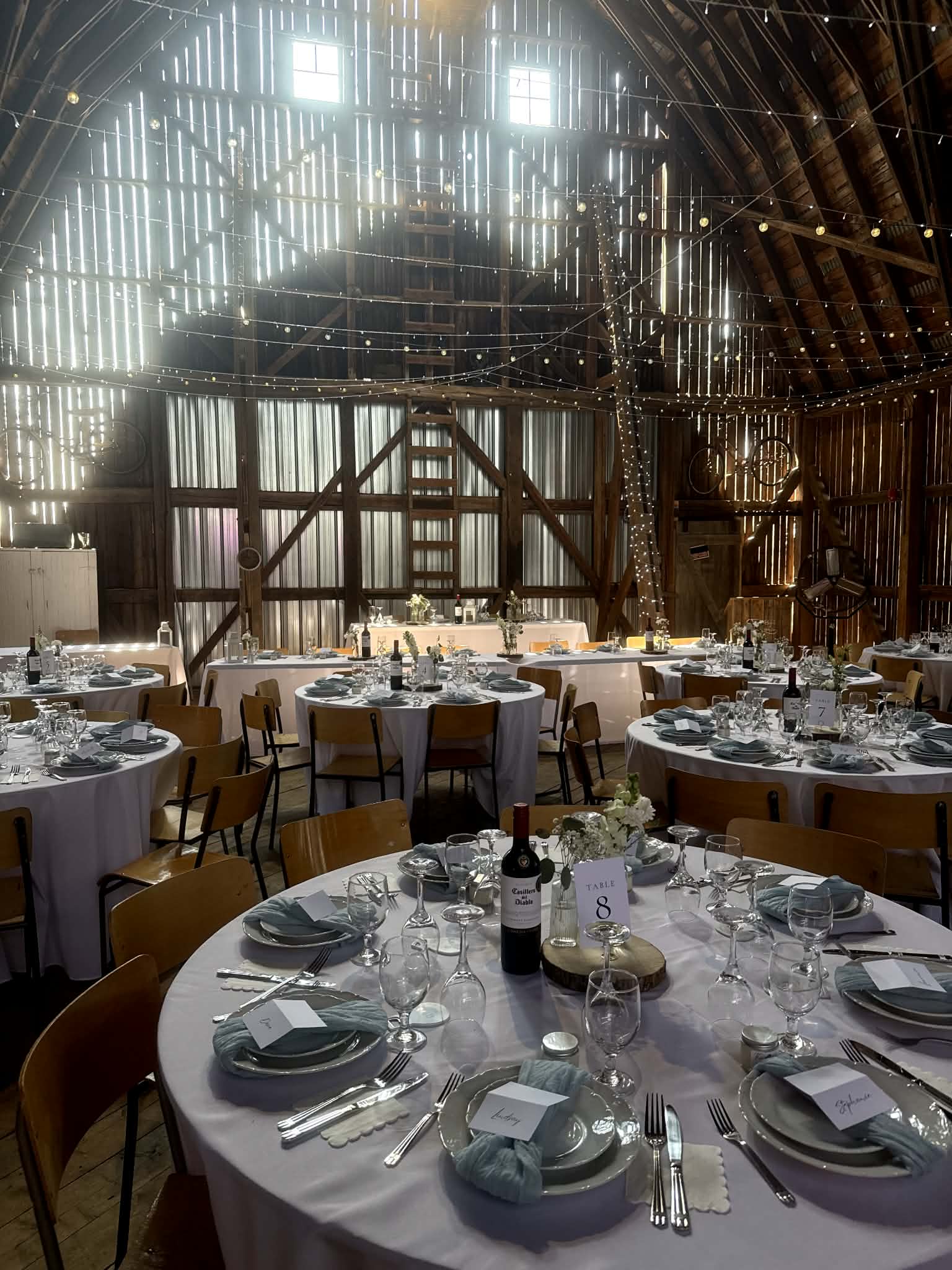Round tables laid with sage napkins and glassware under a canopy of string lights strung between the barn rafters