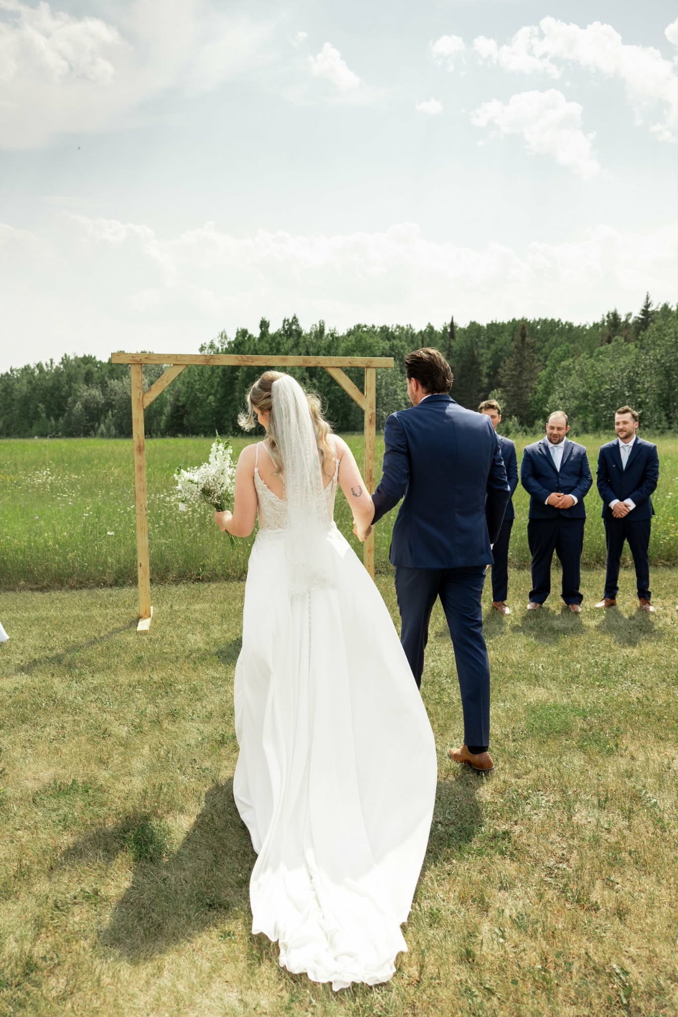 Bride and groom walking hand in hand toward a simple timber arch in the bottom field, groomsmen watching from the treeline
