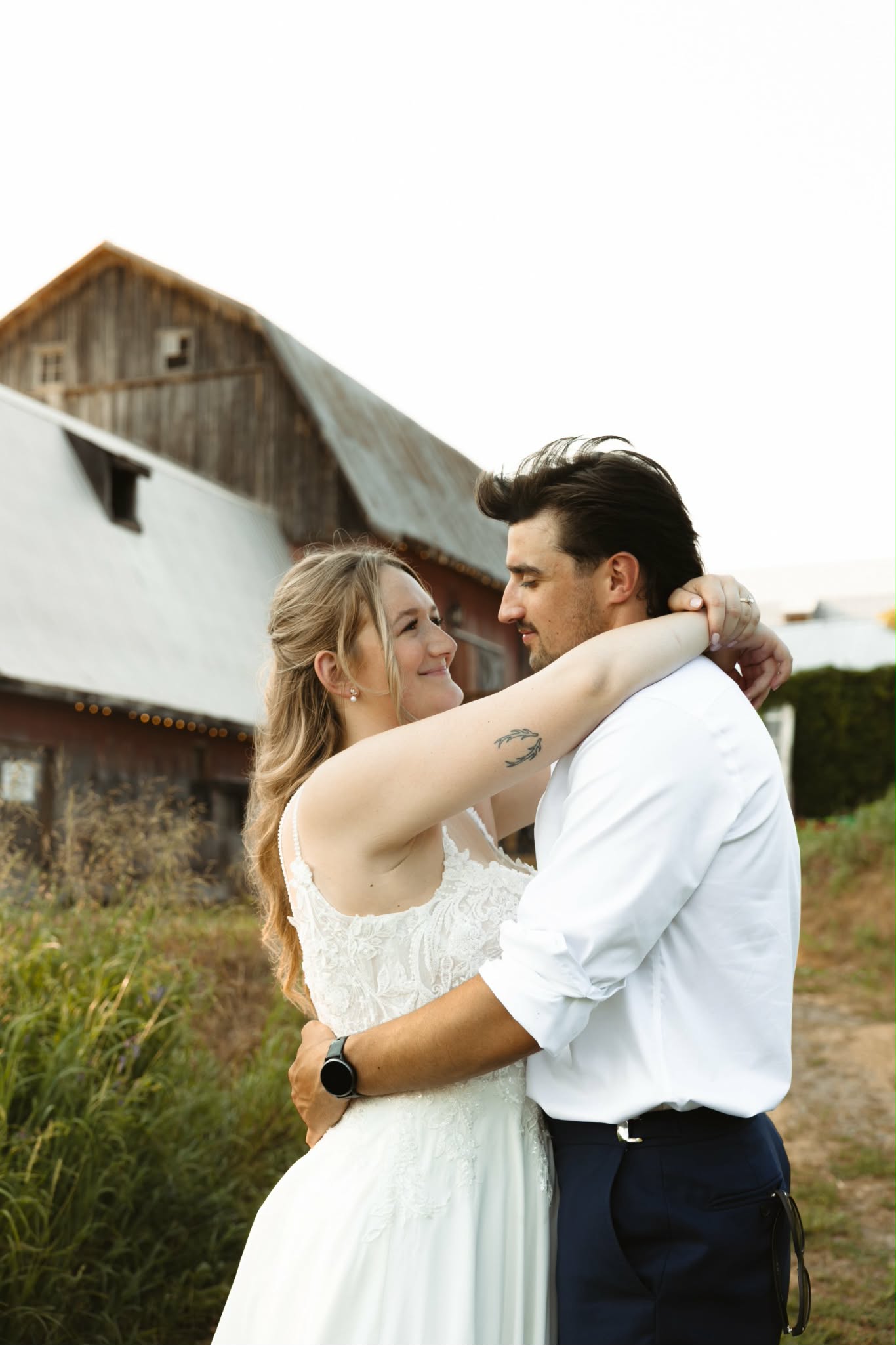 Bride and groom embracing in the long grass at golden hour with the weathered barn behind them