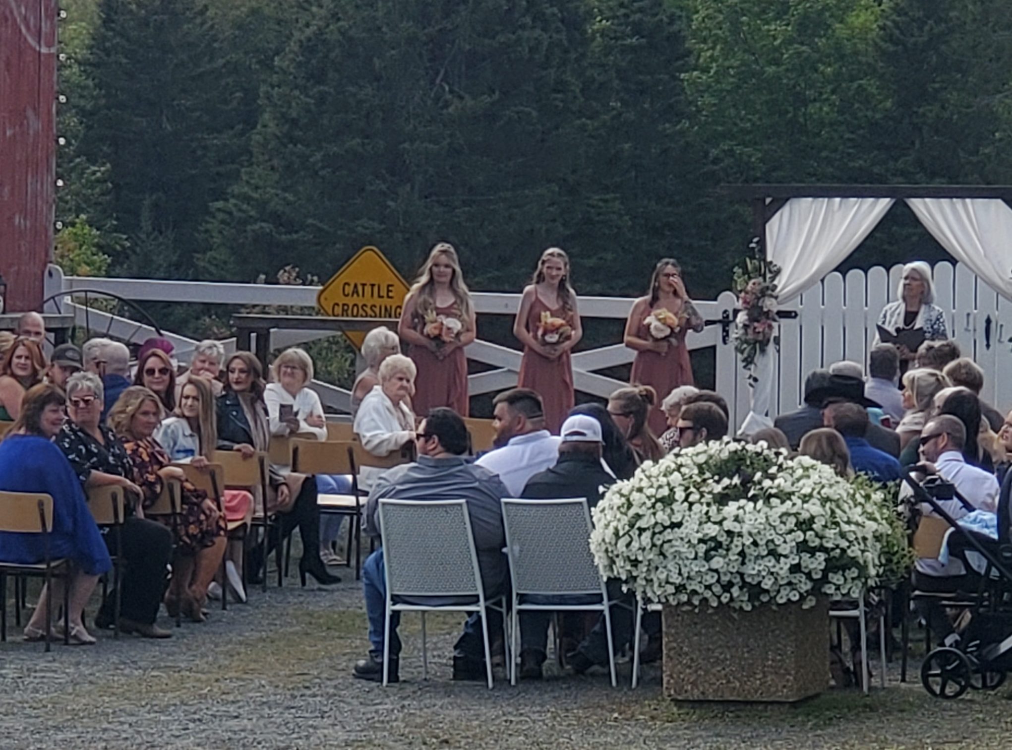 Outdoor ceremony in front of the barn with bridesmaids at a white gate and guests seated in the sunshine